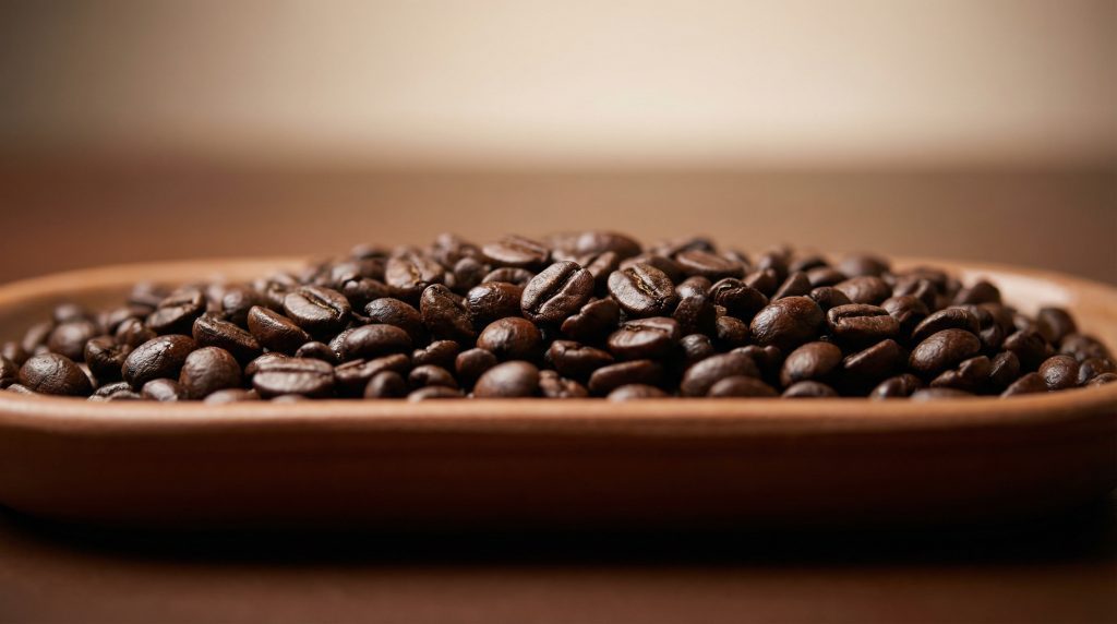 Speciality coffee beans โ close-up texture Create a 16:9 hero banner image that celebrates speciality coffee beans. Straight-on, front-facing view of a shallow ceramic dish or tray filled with freshly roasted coffee beans, seen from directly in front, not from above. The beans are in sharp focus in the centre, with a soft blur towards the edges. Background is a smooth gradient from warm dark brown at the bottom to soft light beige at the top, suggesting depth without distraction. Lighting is gentle and directional from the front, bringing out the oils and texture on the beans. No bags, no packaging, no grinders or machines, no written text or symbols. Photorealistic, high detail, simple and minimal.